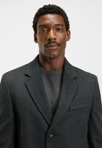 Donkergrijze wolblend blazer met notch revers, enkele knoopsluiting en een borstzak. Ggedragen over een grijze sweater.