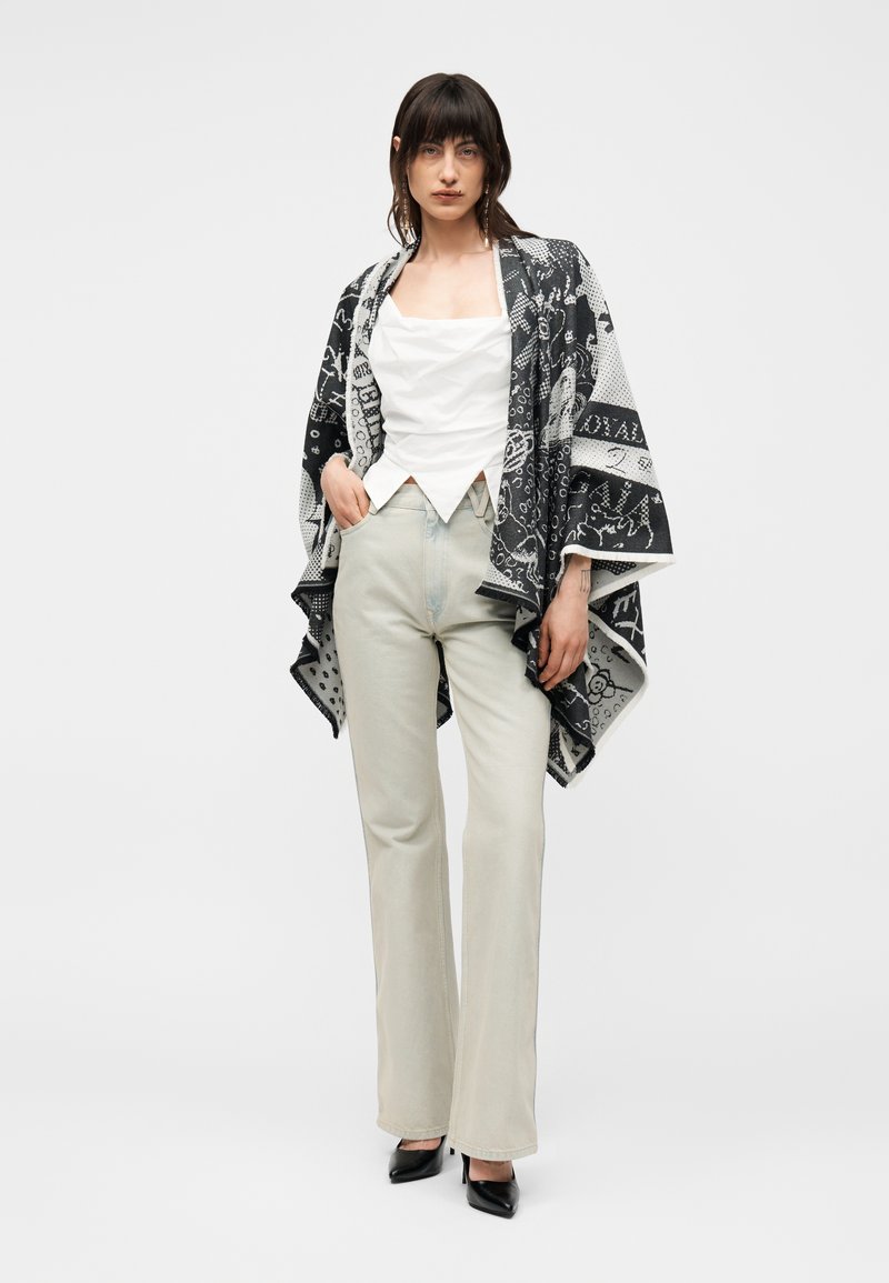 Femme aux cheveux foncés porte un haut blanc sans manches, un pantalon clair taille haute, un châle à motif noir et blanc et des talons noirs pointus, debout.