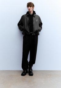 Zwarte bomberjack met rits en logo, gedragen over een zwarte hoodie en broek, gecombineerd met zwarte schoenen, staand tegen een lichte muur.