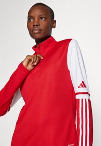 Rød atletisk top med høj krave, lynlås og lange ærmer. Hvide ærmer har rød tre-stribe detalje og logo.