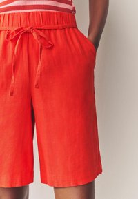 Pantaloni shorts in lino rosso con cintura e lacci regolabili. Presentano tasche laterali e una vestibilità rilassata. Texture liscia e design fino al ginocchio.