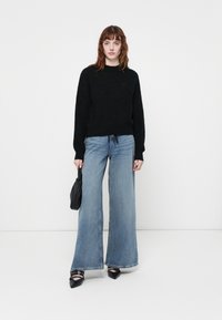 Zwarte gebreide trui met een ronde hals, gecombineerd met lichtblauwe high-waisted wijde jeans. Model houdt een kleine zwarte tas vast.