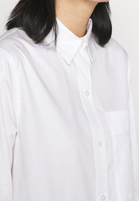 Donna con capelli neri che indossa una camicia bianca con colletto e bottoni, dotata di taschino sul petto, mostrata dal collo fino a metà torso su uno sfondo semplice.