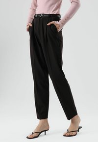 Pantalon noir ajusté taille haute avec plis et poches latérales, associé à un haut à manches longues rose clair et des sandales à talons aiguilles noires.