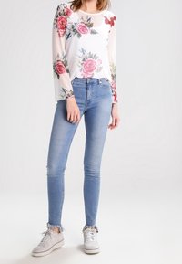 Průsvitný bílý top s květinovým potiskem, dlouhými rukávy, v kombinaci s světle modrými těsnými džínami a šedými teniskami s texturovanými detaily.