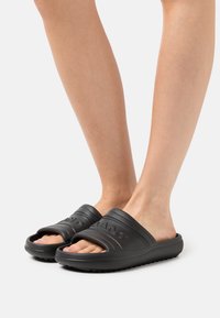 Black slide sandals with a textured rubber sole, featuring a wide band across the top embossed with the brand name.