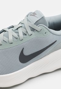 Baskets de sport gris clair avec une tige en maille texturée, logo swoosh gris foncé et semelle en caoutchouc blanche. Comprend des lacets plats et une languette rembourrée.