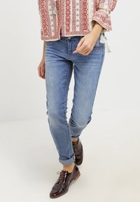 Jeans de mezclilla azul claro con un corte ajustado, puños enrollados y desvanecimiento sutil, combinados con zapatos de cuero marrón oscuro.