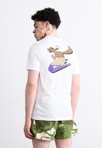 Witte katoenen T-shirt met een cartoonkangoeroe en het "NIKE" logo in paars op de achterkant; gecombineerd met groene met patronen bedrukte shorts.