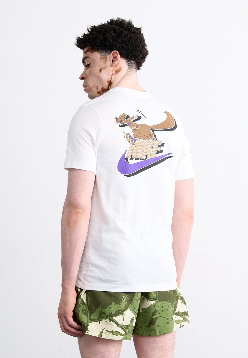 Witte katoenen T-shirt met een cartoonkangoeroe en het "NIKE" logo in paars op de achterkant; gecombineerd met groene met patronen bedrukte shorts.