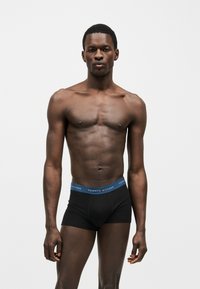 Άνδρας μοντέλο με σκούρο δέρμα και κοντά μαλλιά, φοράει μαύρα εσώρουχα Tommy Hilfiger με μπλε μέση, σε απλό λευκό φόντο.