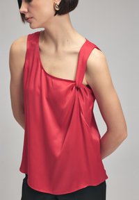 Rød satin top med en enkelt skulderstrop, der har en knude detalje på den ene side og et flydende, draperet design. Glat tekstur og rund kant.