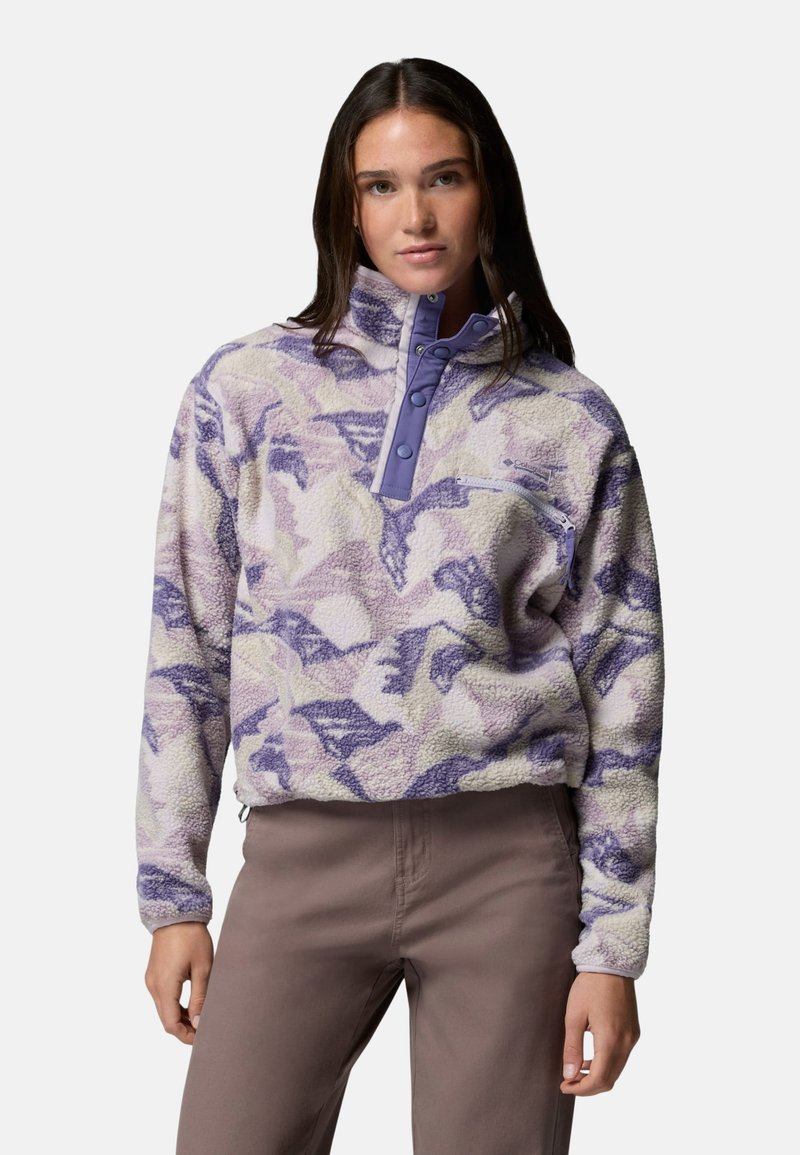Femme portant un pull polaire camouflage violet et beige avec des boutons-pression et une poche poitrine zippée, associé à un pantalon marron.
