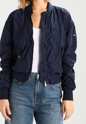 Woman wearing a navy blue bomber jacket over a white tank top and blue jeans, standing against a plain light gray background.