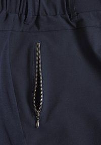 Primo piano di pantaloni navy con cerniera verticale argentata parzialmente aperta e vita elastica con passanti per cintura.
