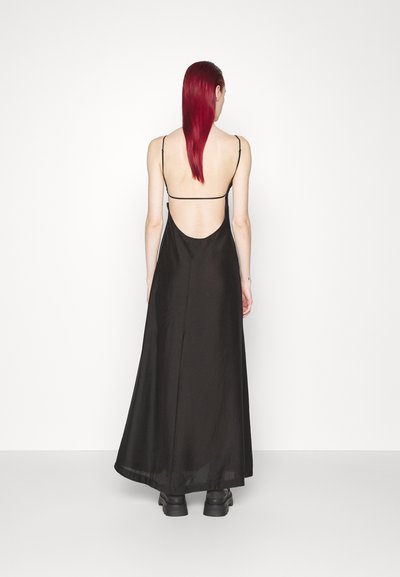 Čierna saténová maxi šaty s tenkými ramienkami, otvoreným chrbtom, voľným strihom a hladkou textúrou. Kombinované s robustnou čiernou obuvou.
