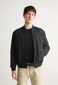 Veste bomber noire avec fermeture éclair à l'avant, col et poignets côtelés. Tissu léger, texture légèrement froissée, poches latérales et détails subtils.
