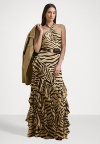 Pitkä halterneck-mekko, jossa on tiikeriraidoitus beige- ja ruskeansävyissä, ja jossa on röyhelökerroksia. Mekko on yhdistetty ruskeaan vyöhön ja vihreään takkiin.