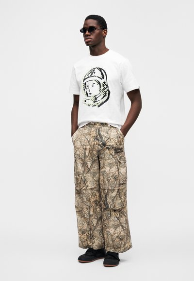 Λευκό μπλουζάκι με γραφικό αστροναύτη, συνδυασμένο με μπεζ cargo παντελόνια καμουφλάζ με πλαϊνές τσέπες. Μαύρα παπούτσια slip-on ολοκληρώνουν την εμφάνιση.