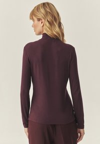 Hosszú ujjú, bordó felső, magas gallérral, puha anyagból készült; testhezálló fazonú, diszkrét fényű. Nincsenek látható minták vagy fémalkatrészek.