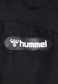 Czarna tkanina z dużą czarno-białą metką logo, na której widnieje napis "hummel". Materiał wygląda gładko i ma lekki połysk.