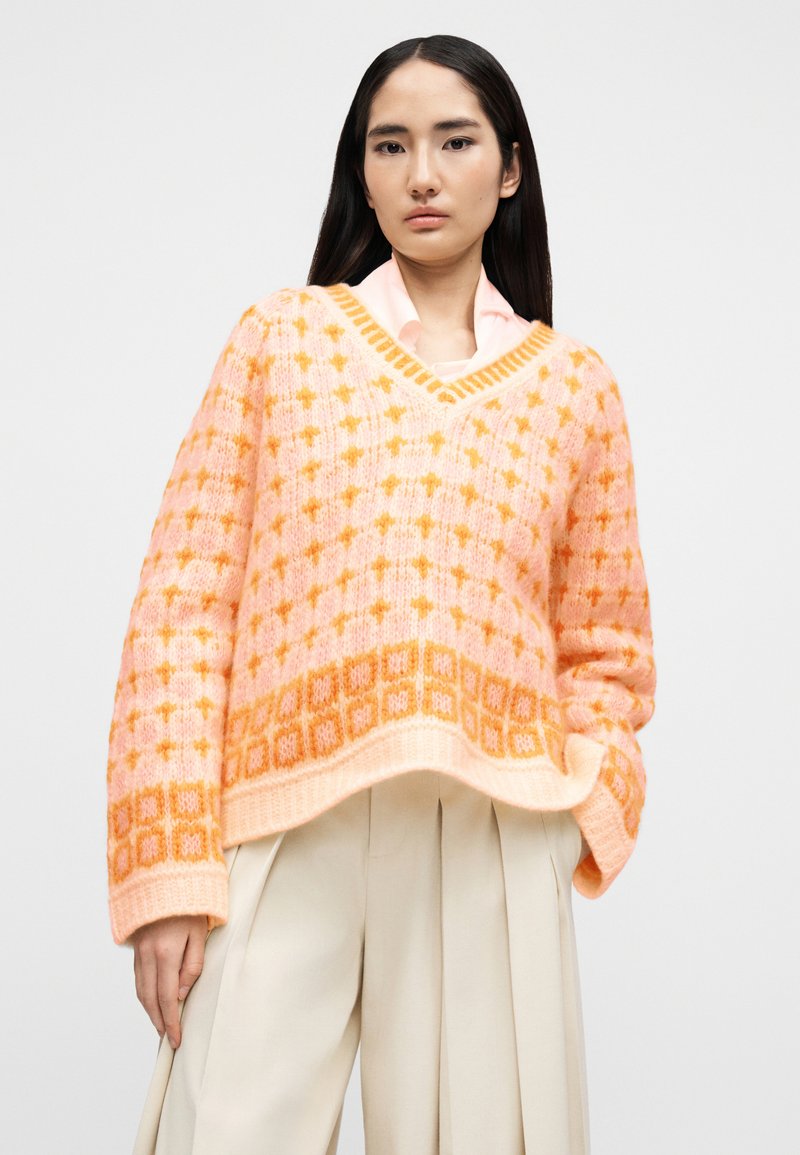 Femme portant un pull en maille à motifs roses et oranges par-dessus une chemise rose pâle et un pantalon large crème, debout devant un fond uni.