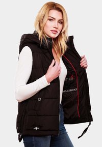 Gilet rembourré noir avec capuche, design matelassé et zips visibles. L'intérieur présente des accents rouges et des poches de rangement. Porté sur un haut blanc.