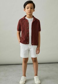 Cárdigan de punto burdeos de manga corta con patrón de diamantes, camiseta blanca, pantalones cortos blancos y zapatillas blancas, de pie sobre un fondo neutro.