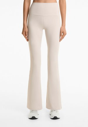 Cream-colored flared leggings made of smooth, stretchy fabric, featuring a high waistband and a full-length design, paired with white sneakers.