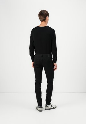 Jeune personne debout de dos, portant un t-shirt noir à manches longues, un jean slim noir et des baskets blanches avec des accents noirs.