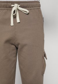 Brown sweatpants with a soft texture, elastic waistband, and cream drawstring. Features side pockets and a short zipper detail.