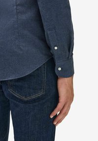 Mörkblå långärmad skjorta med knappar i en texturerad tyg, med knappmanschetter och sidoklyfta, ihop med mörk denimjeans.