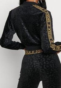 Tuta in velluto nero con motivo leopardato, dotata di collo e polsini a costine. Accenti distintivi in oro sul logo presenti sulle maniche e sulla cintura.