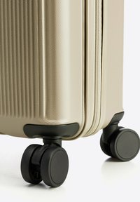 Tuhý kufr v svetle béžové barvě, s texturovaným povrchem, dvojitými černými kolečky a hladkým zipem po boku.