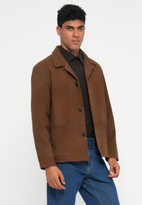 Brown cotton jacket with a collared neck, button closure, two front pockets, and a relaxed fit, paired with a black shirt and blue jeans.