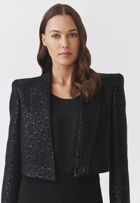 Kurz geschnittene schwarze Jacke aus strukturiertem, gemustertem Stoff, mit Schulterpolstern und einem einzigen Knopfverschluss. Wird über einem schwarzen Top getragen.