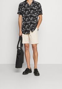 Musta lyhythihainen paita, jossa valkoinen palmukuvio, kermahilloset shortsit, mustat kengät ja musta tikattu kangaskassi. Yksinkertainen muotoilu, rento ilme.