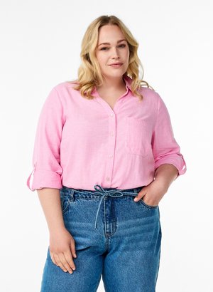 Lyshåret kvinde med krøllet hår iført en pink knappet skjorte med opklemte ærmer og højtaljede blå jeans, den ene hånd i lommen.