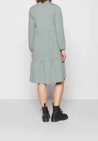 Robe verte à motifs avec des manches longues, un design à volants et un col rond, associée à des bottines noires à enfiler avec des accents jaunes.