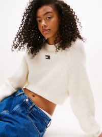 Cream-colored ribbed knit sweater with a cropped design, featuring a small logo patch, paired with high-waisted blue denim jeans.