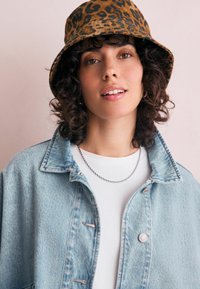Chapeau seau à motifs léopard marron, veste en denim bleu clair, t-shirt blanc, collier en chaîne argentée, présentant une texture douce, une forme ronde et des coutures visibles.