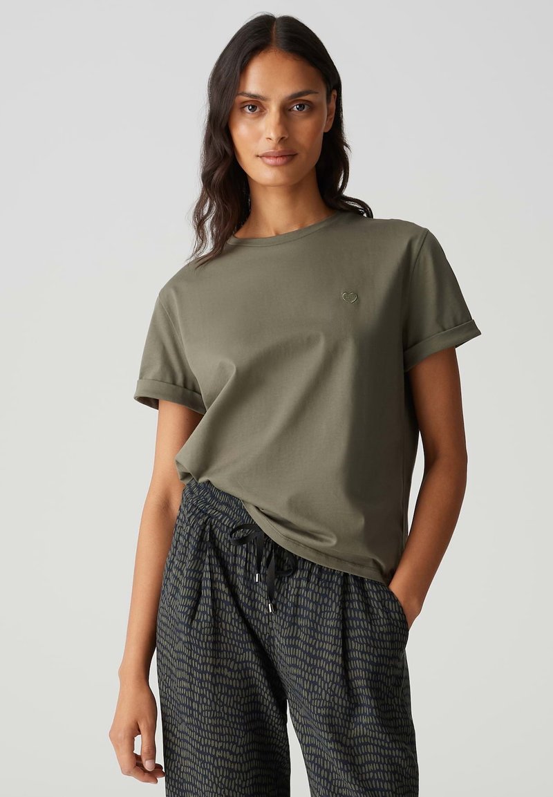 T-shirt en coton vert olive avec des manches courtes roulées et un petit logo en forme de cœur sur la poitrine gauche, associé à un pantalon noir à motifs.