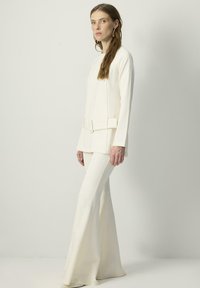 Kreemikaalane kohandatud kost�üüm, mis sisaldab pikka varrukaga pluusi, millel on vöökoht ja laiad püksid. Läikiv materjal, millel on sile tekstuur.