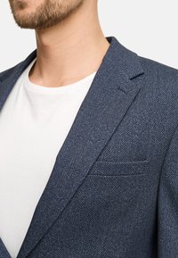 Marine herringbone blazer van textiel met structuur, met notch revers en een getailleerde fit. Draag het over een witte t-shirt, waarbij een deel van de kraag en het zakdetail zichtbaar zijn.