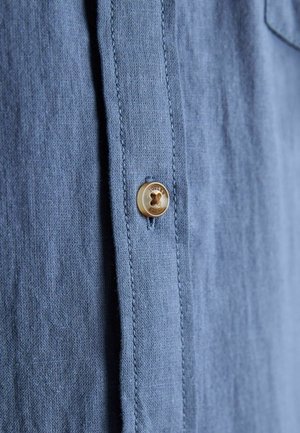 Prim-plan al unei cămăși din in bleu deschis, arătând un nasture bej cu patru găuri cusut pe capse.