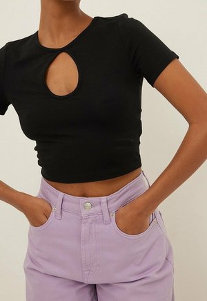 Kobieta ubrana w dopasowany czarny krótki top z wycięciem w kształcie łzy na wysokości piersi oraz lawendowe spodnie z wysokim stanem, mająca ręce w kieszeniach.