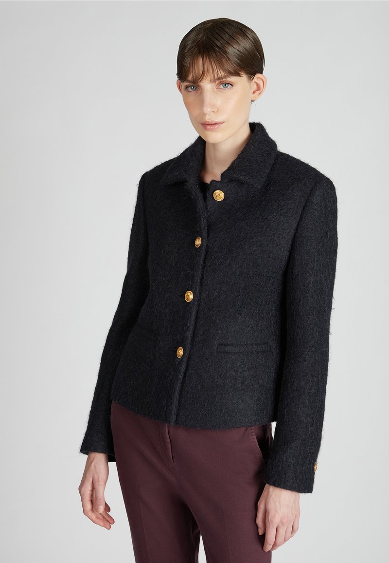Chaqueta de lana negra con cuello, adornada con botones dorados y bolsillos laterales. La tela tiene textura, creando una silueta definida.