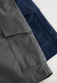Shorts cargo gris et bleu marine pliés, mettant en valeur de grandes poches latérales avec rabats et une double couture visible sur un fond clair.