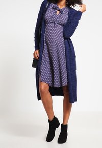 En marinblå stickad tröja över en blommönstrad klänning med en rosett vid halsen, matchad med svarta ankelskor. Klänningen har korta ärmar och en knälång längd.