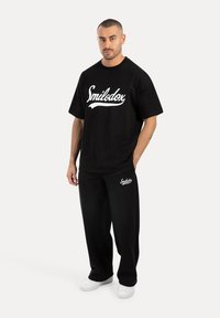 Svart bomullströja med vit "Smilodox"-logotyp, ihop med matchande svarta byxor. Stiliserat med vita sneakers och avslappnad passform.